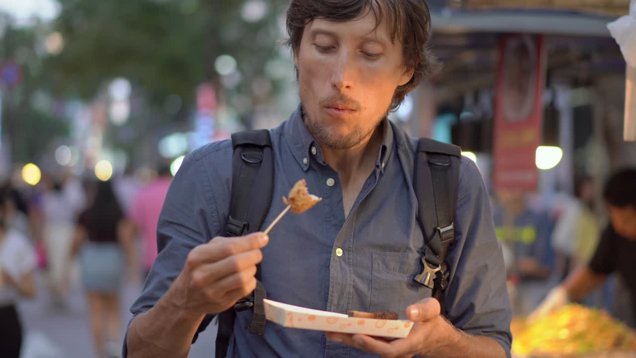 un joven visita el tradicional mercado callejero coreano. está comiendo pastel de pescado en un palo de bambú. viaje a corea