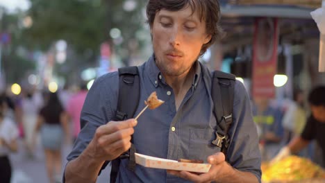 young man visits the traditional korean street market. he is eating fish cake on a bamboo stick. travel to korea