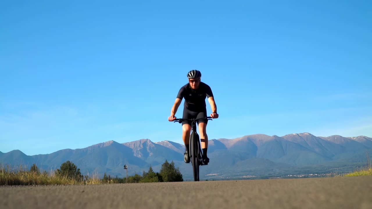 Un singolo ciclista pedala lungo un'ampia strada rurale con montagne distanti, campi aperti e un cielo azzurro limpido in piena luce diurna, creando un senso di viaggio.