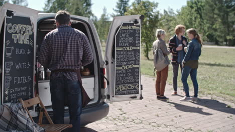 coffee truck worker leaning on the van while using a tablet, group of people talk and drink coffee in the background