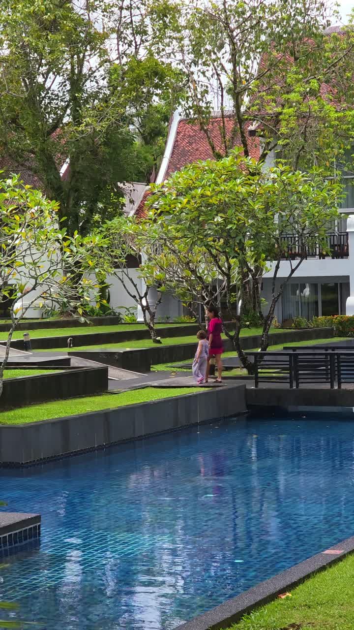 Family strolls along sparkling blue pool surrounded by green lawns and flowering trees in an elegant vacation setting in Thailand
