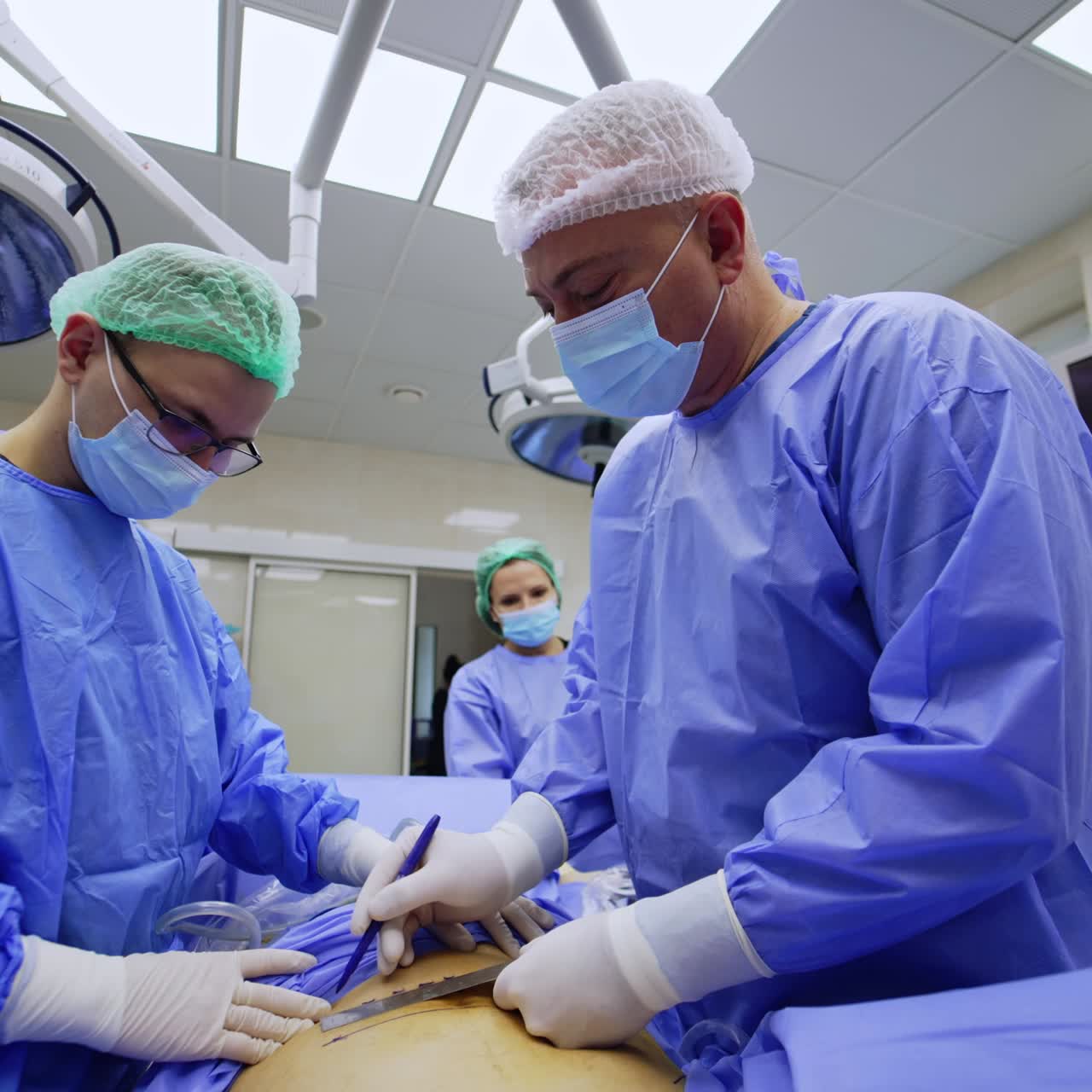 Two male surgeons prepare the patient for procedure. Older doctor applies lines with marker pen and metal ruler. Low angle view