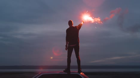 handheld cinematic dark shot of young man standing on the car with red signal burning flare on the beach near the water