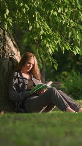 mujer sentada al aire libre con las piernas estiradas, apoyada en un árbol en un campo de hierba, leyendo un libro bajo la cálida luz del sol, las hojas de los árboles se balancean suavemente en la brisa, el fondo presenta un edificio y vegetación