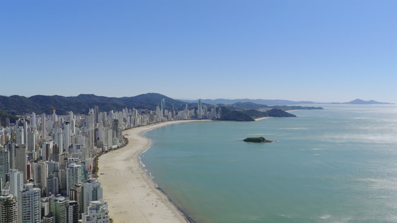 Balneario Camboriu urban landscape revealing sandy shoreline, azure waters, mountain silhouettes during golden sunlight, showcasing Brazilian coastal panorama from aerial drone perspective