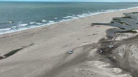 aerial: white suv on jericoacoara beach with atlantic ocean during the day in jijoca de jericoacoara, ceara, brazil, tracking drone shot