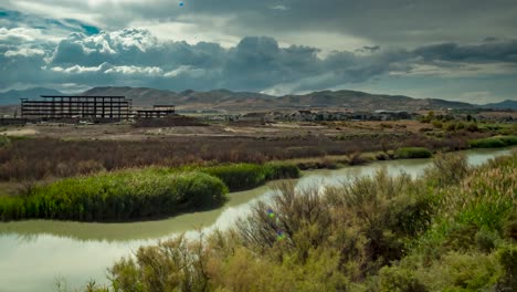 Cloudscape-above-a-river,-mountains-and-a-construction-site---panning-time-lapse