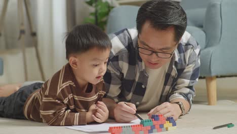 close up of asian father and son lying on the floor in the room with plastic toy brick drawing together at home