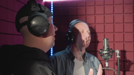 two men in a soundproof recording studio. one man in a black cap and hoodie smiles while watching the other, dressed in a blue shirt and white undershirt, singing passionately