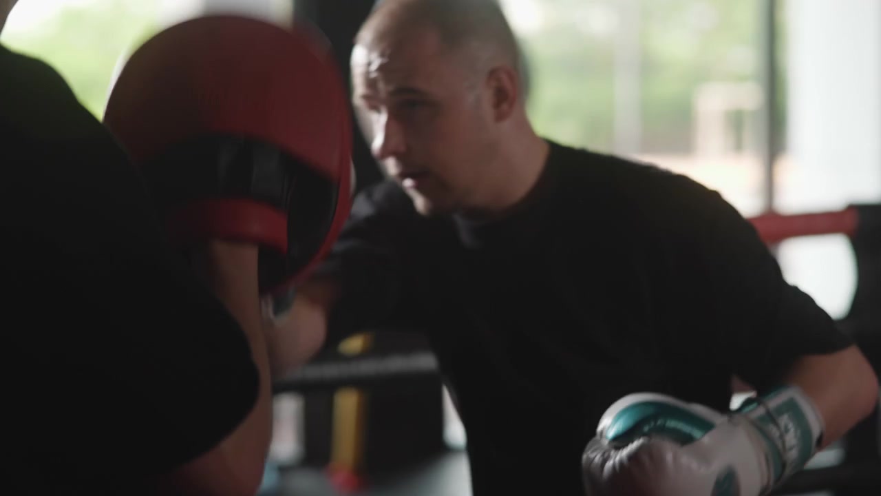 Two men train inside boxing ring as coach braces red focus mitt and boxer with gloved hands delivers sharp punches in intense practice, framed by window backlight and brick walls, with boxing