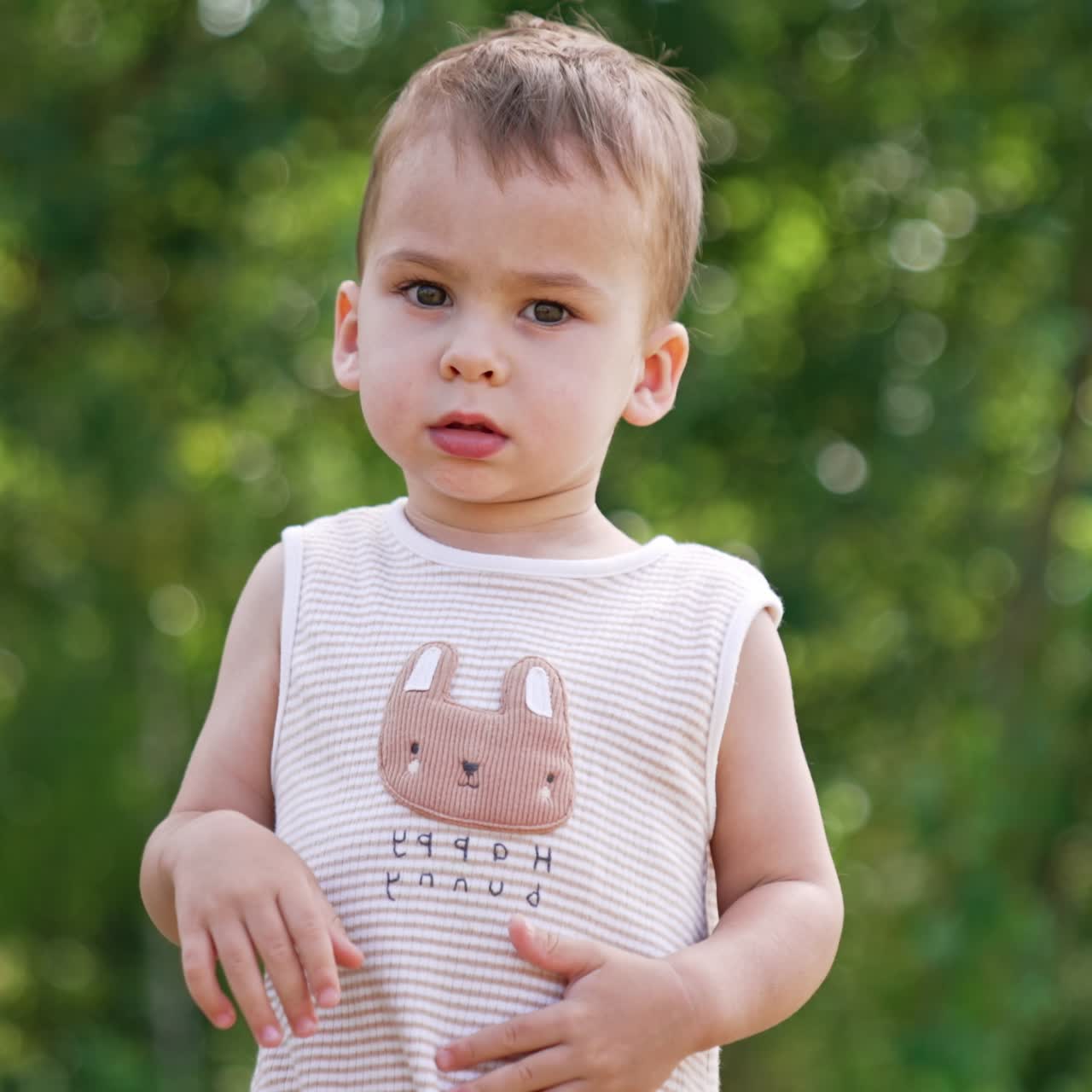 Beautiful toddler says something gesturing with hands. Kid waves his hands and looks at camera. Blurred greenery of nature at backdrop
