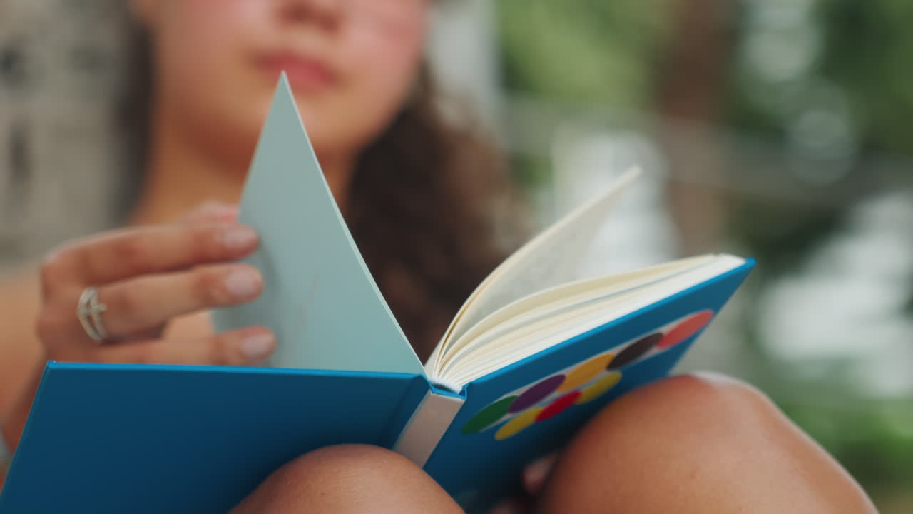 Giovane donna che legge un quaderno blu sulle ginocchia all'aperto, primo piano delle mani che girano le pagine, sole pomeridiano, palme del resort, scatto di vita spontaneo che mostra anelli e manicure, le scene suggeriscono studentessa, scrittrice
