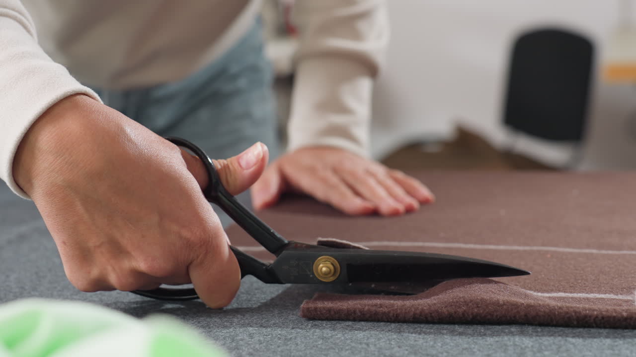 Fashion designer using large scissors cutting thick brown material along chalk markings on worktable, wearing casual light sweater and jeans, carefully preparing fabric pieces for sewing project