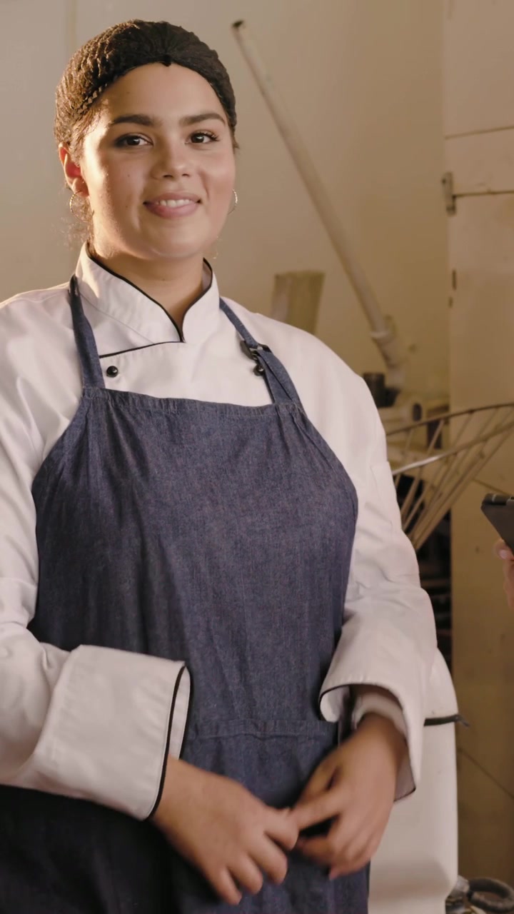 Vídeo vertical: Chef gesticulando de jaleco apressando mulher verificando tablet perto dos fornos confirmando pedido