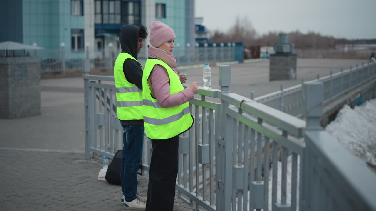 Two people in reflective safety vests stand by riverside railing in cold weather near urban buildings, looking toward distant walkway and partially frozen river, with backpack and bags placed on ground