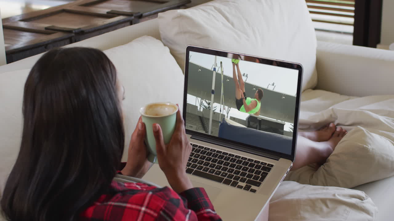 Composite of woman sitting at home holding coffee watching athletics high jump event on laptop