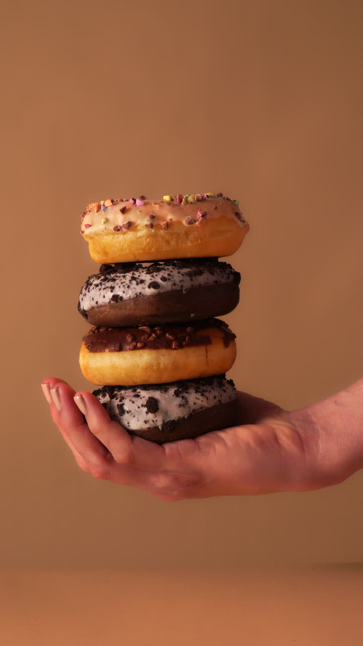 Close up of hands stacking the final vanilla glazed donut with colorful sprinkles on top of chocolate iced and cookie crumb donuts. Warm beige studio background with clean minimal look