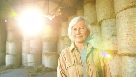 retrato de una anciana campesina caucásica de pelo gris parada en un establo con existencias de heno y sonriendo a la cámara