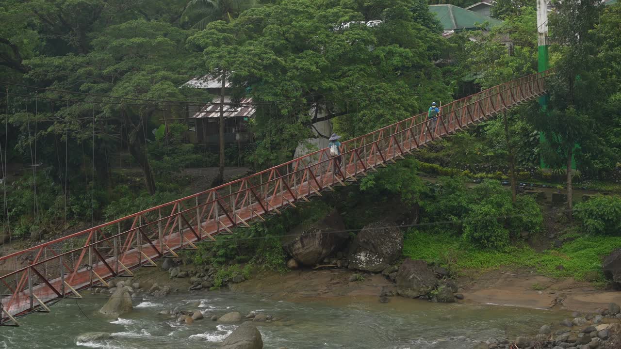 Framed from a forest-side angle, two people cross a red suspension bridge over a steady Philippine river, surrounded by lush greenery and distant buildings nestled in trees