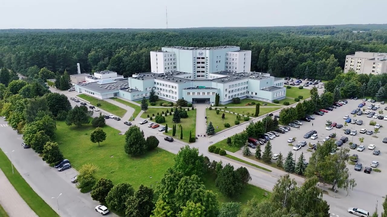 Nemocnice námořníků Klaipėda se tyčí nad okolními parkovišti a trávníky, zatímco dron letí nad upraveným lékařským kampusem.