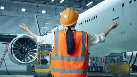 vista trasera de una ingeniera asiática con casco de seguridad extendiendo los brazos mientras está de pie con el avión en el hangar