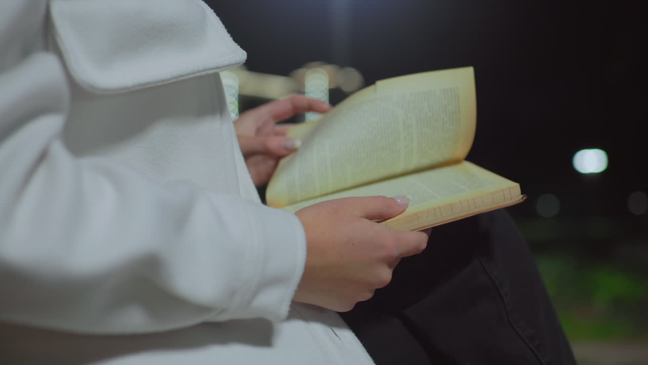 Close up of girl wearing white coat with polished nails gently flipping page of book under bright light at night with soft bokeh effect in background
