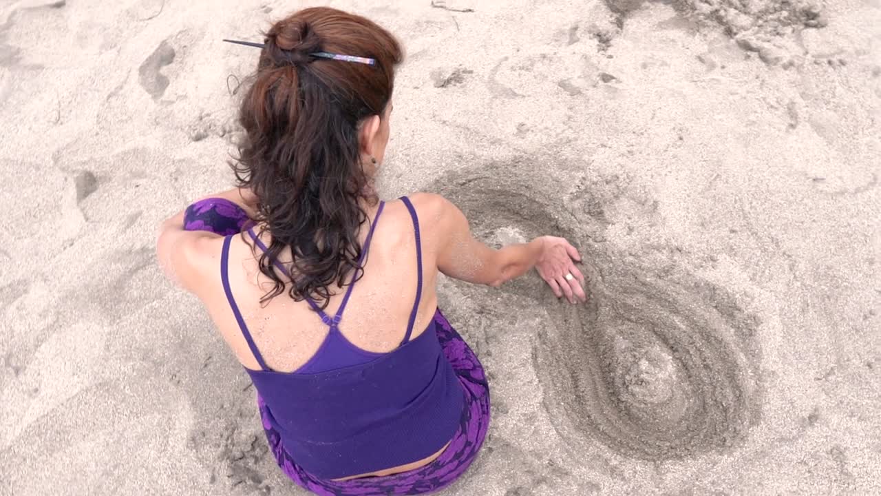 A woman dressed in striking purple is seen from above as she meticulously crafts an infinity symbol in the sand