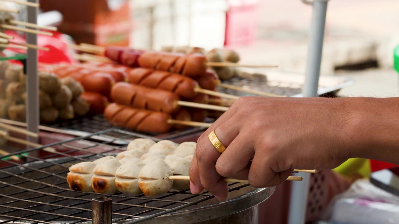 Close-up tangan penjual membalik bakso dan sosis bakar di Pasar Bunga Bangkok