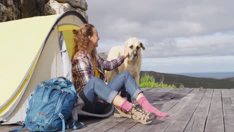 happy caucasian woman camping sitting outside tent with pet dog, putting on boots on mountainside