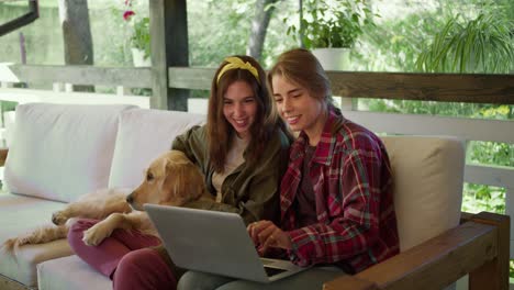 a blonde girl in a red plaid shirt and a brunette girl in a green t-shirt are sitting and looking at something on a laptop, a dog is sitting next to them, they are in a gazebo in nature