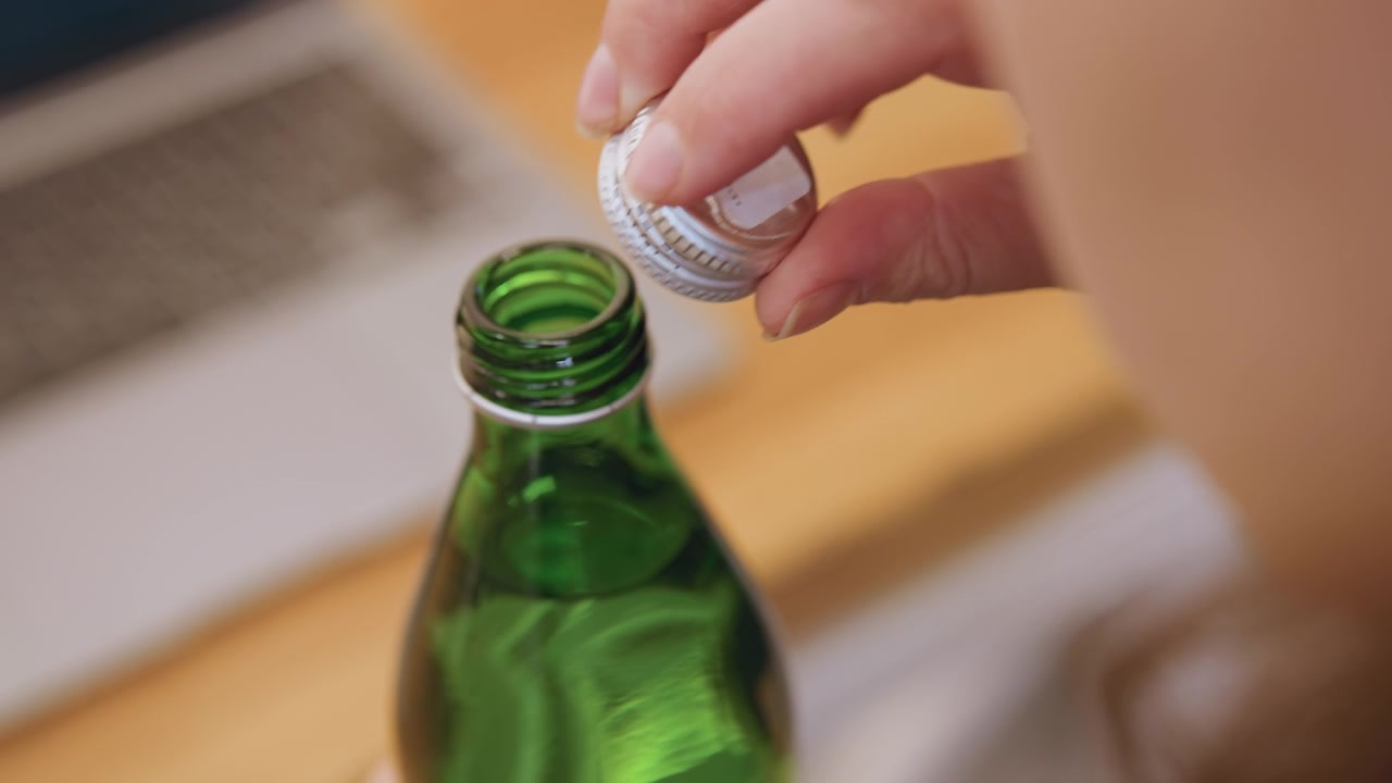 Bouteille verte avec un bouchon en plastique blanc, gros plan sur des mains retirant le bouchon sur un bureau en bois près d'un ordinateur portable, un créateur de contenu effectuant un petit rituel pour ouvrir une boisson, détail du filetage tactile et espace de travail calme.