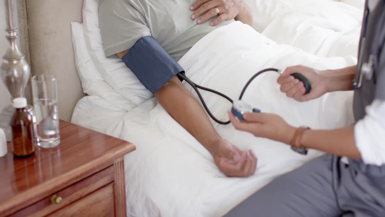 Medico uomo multietnico che misura la pressione sanguigna a paziente anziano uomo a letto a casa, al rallentatore