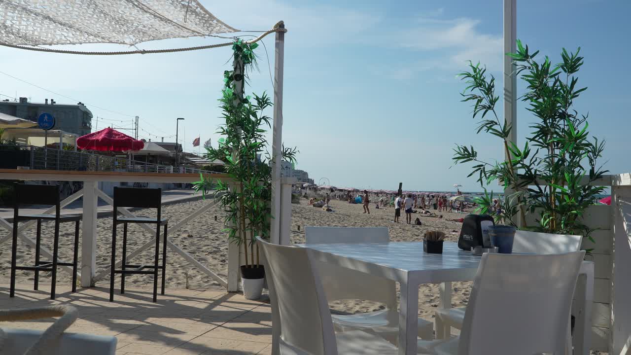 A scenic view from a beach café overlooking the sandy beach, with children playing nearby. The scene captures the relaxed atmosphere of a sunny day at the beach, perfect for leisure and relaxation.