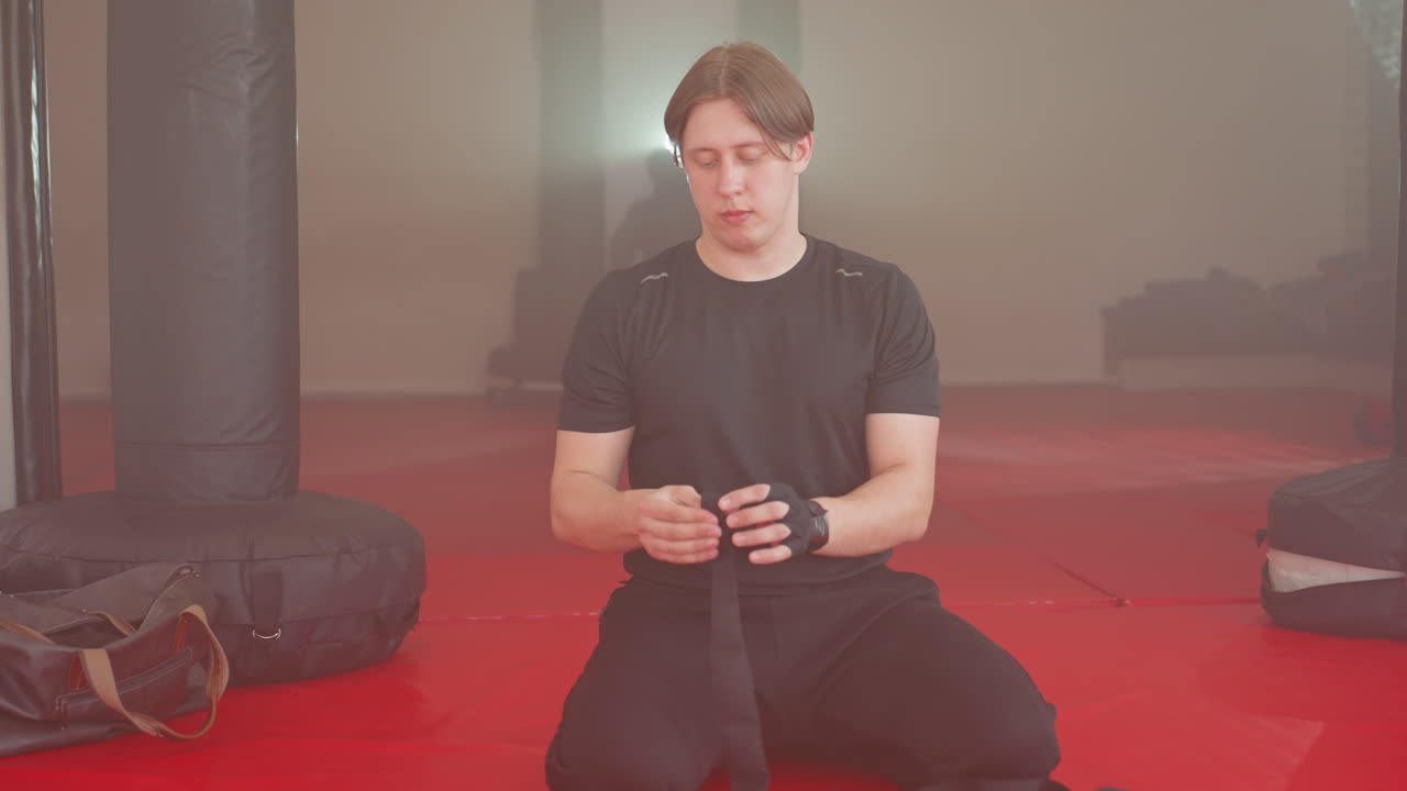 Boxer in black sportswear sits on red mat gym floor wrapping long hand bandages with focus and precision, preparing for training session with punching bags around, showing strength and readiness for combat