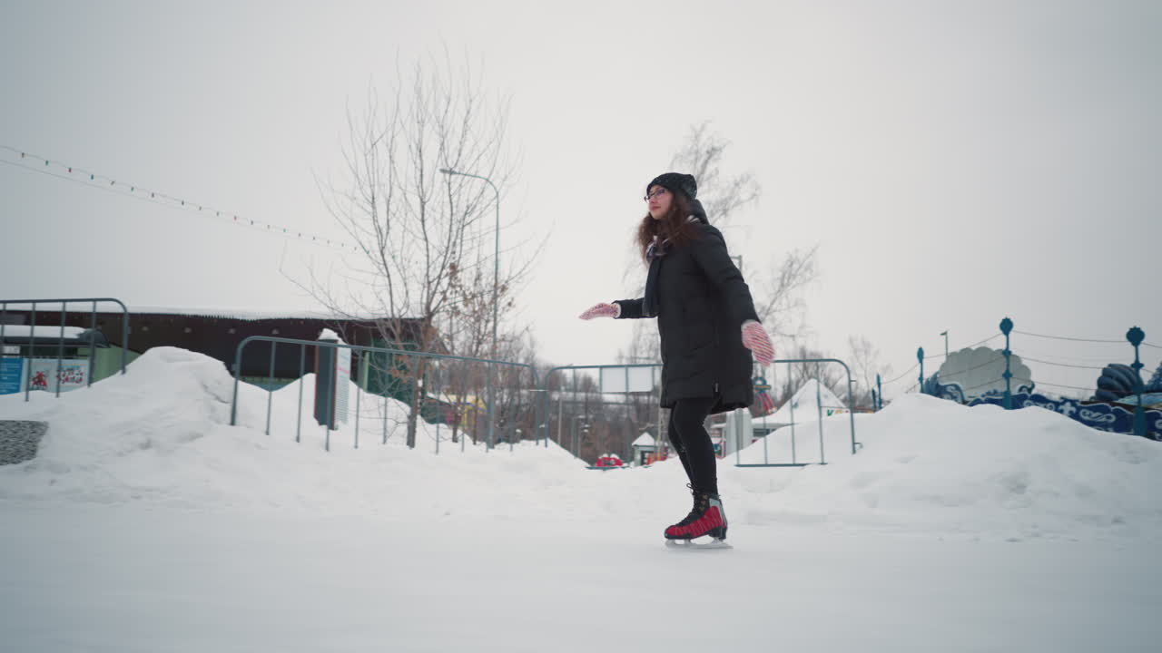 Lady skating gracefully on outdoor ice rink during winter, dressed in black coat, red skates, scarf, and warm gloves, surrounded by snow, benches, and decorative park structures under cloudy sky