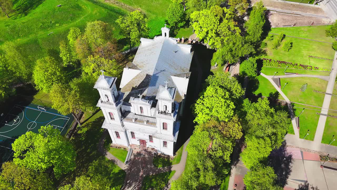 A white historic church with twin towers sits in the middle of a vibrant green park, crisscrossed by paths and surrounded by lush spring trees.