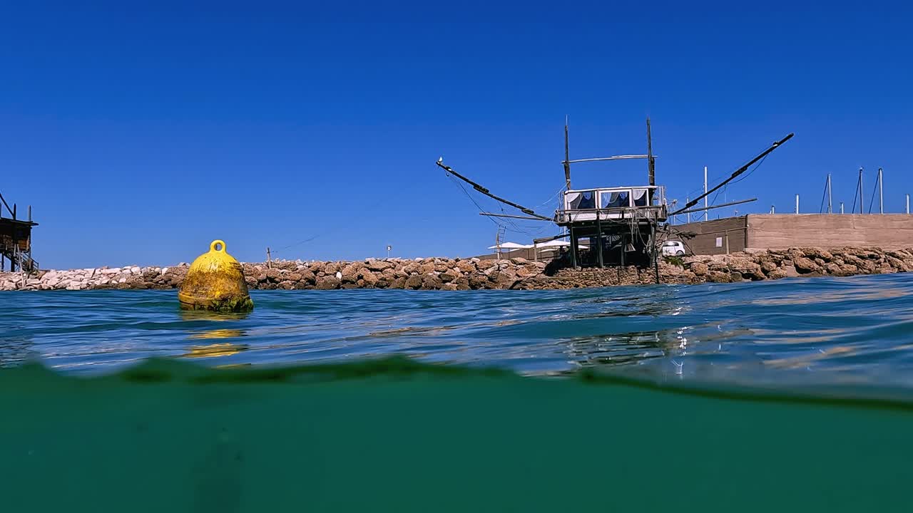 배경에 있는 바위 방파제에 있는 노란색 떠 있는 부표와 트라보치 나무 낚시 플랫폼의 수중 패닝 절반