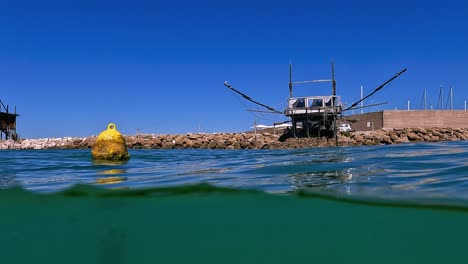 Halbe-Unterwasserschwenkansicht-Der-Gelben-Schwimmenden-Boje-Und-Trabocchi-Fischerplattform-Aus-Holz-Auf-Felsigem-Wellenbrecher-Im-Hintergrund