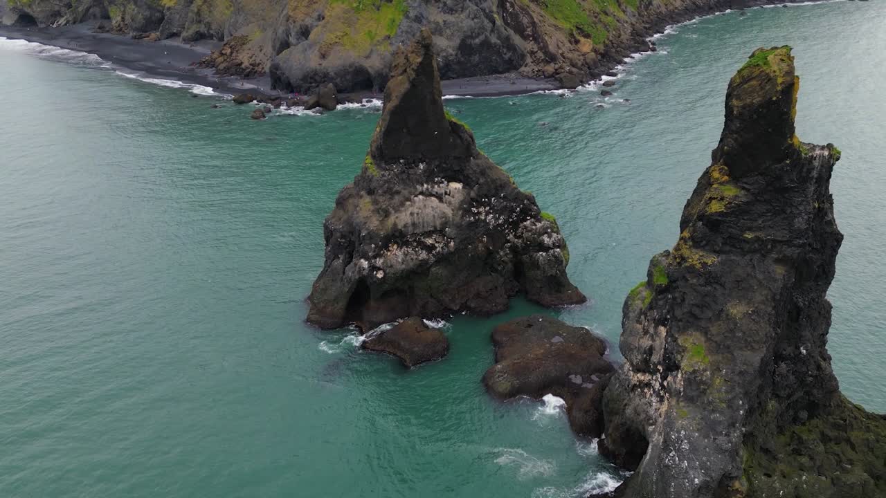 Cinematický záběr dronem ve 4K nad pláží Reynisfjara, zachycující kontrast mezi tmavým sopečným pobřežím, tyrkysovými vlnami a dramatickými útesy pod náladovou severskou oblohou – Iceland_35