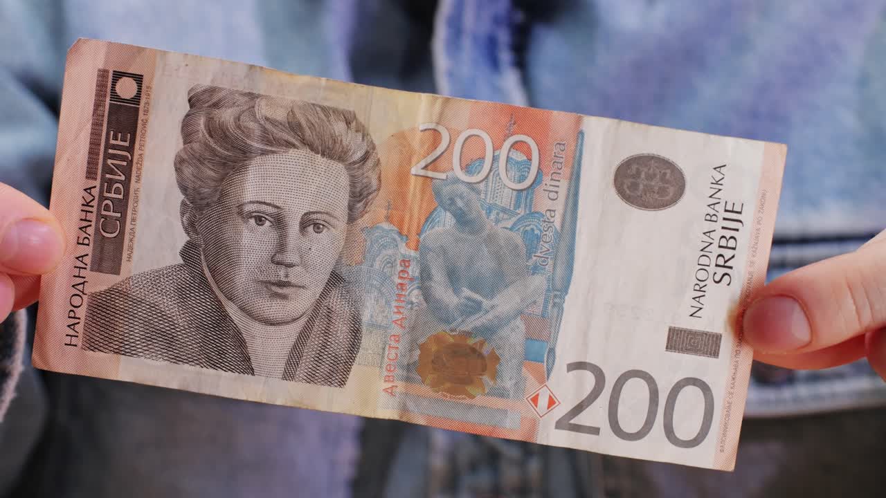 A person is holding a 200 SRB banknote in their hand. The currency is being handled between thumb and fingers, observing the intricate font and art on the paper cash