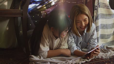 asian mother and daughter smiling while using digital tablet under blanket fort at home