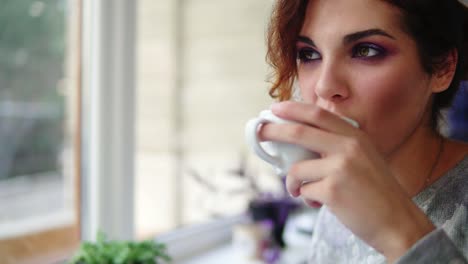 Hermosa-Joven-Disfrutando-De-Un-Café-Y-Sonriendo-Sentada-Junto-A-La-Ventana-En-La-Cafetería.-Señora-Con-Una-Taza-De-Bebida-Humeante-Sonriendo