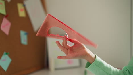 Little-girl-spins-pink-square-ruler-in-fingers-near-corkboard