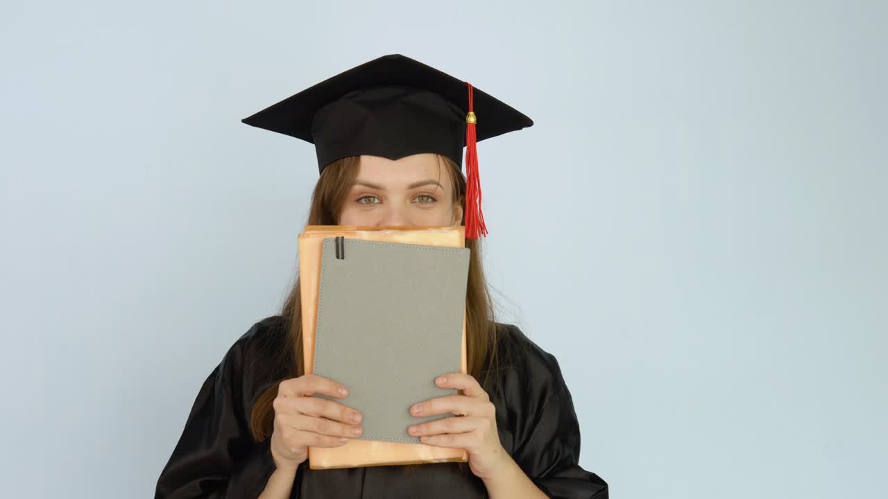 una joven estudiante con una túnica negra y un sombrero de maestro y cubre la mitad inferior de su cara, boca y nariz con libros que sostiene en sus manos.