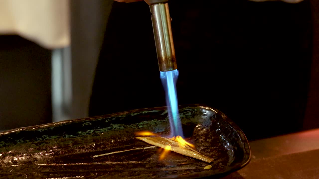 A chef uses a blowtorch to sear anago eel on a black plate, close-up shot
