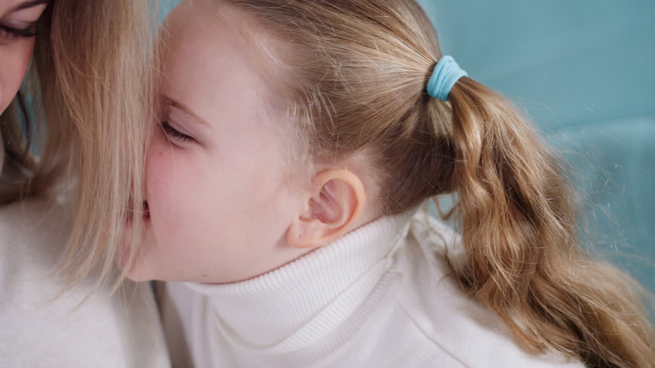 Smiling young girl with blonde hair in ponytail wearing white sweater looks joyful while sitting on turquoise couch, radiating happiness, innocence, carefree childhood spirit genuine candid emotion