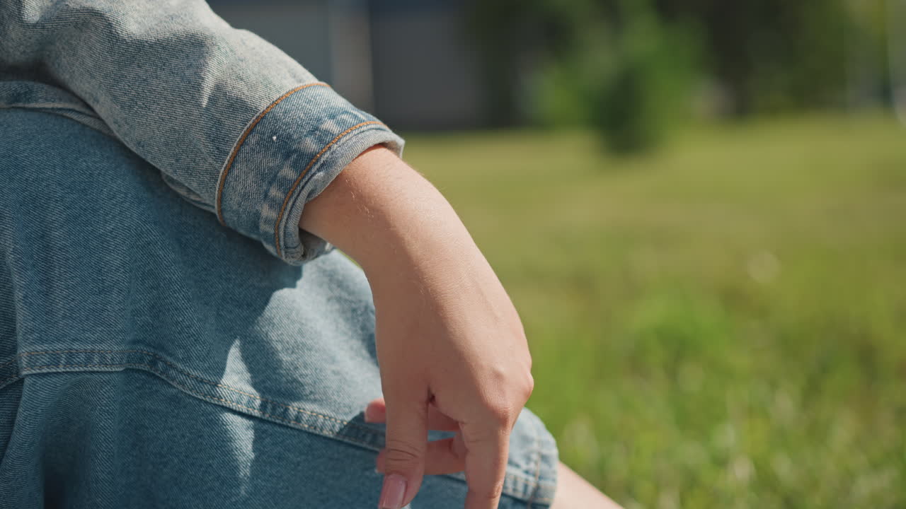 En hand sträcker sig genom gräset i en park, med synlig denimärm och jeans. Fingrarna sprids i en livlig rörelse under starkt sommarljus. Grön äng i bakgrunden, en student skissar i närheten. Lekfull och taktil känsla.