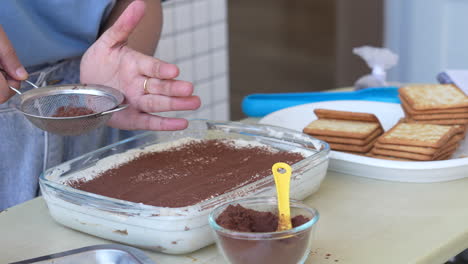 Frauenhand-Streut-Kakao-Auf-Tiramisu-Kuchen-In-Glasschüssel-In-Küche