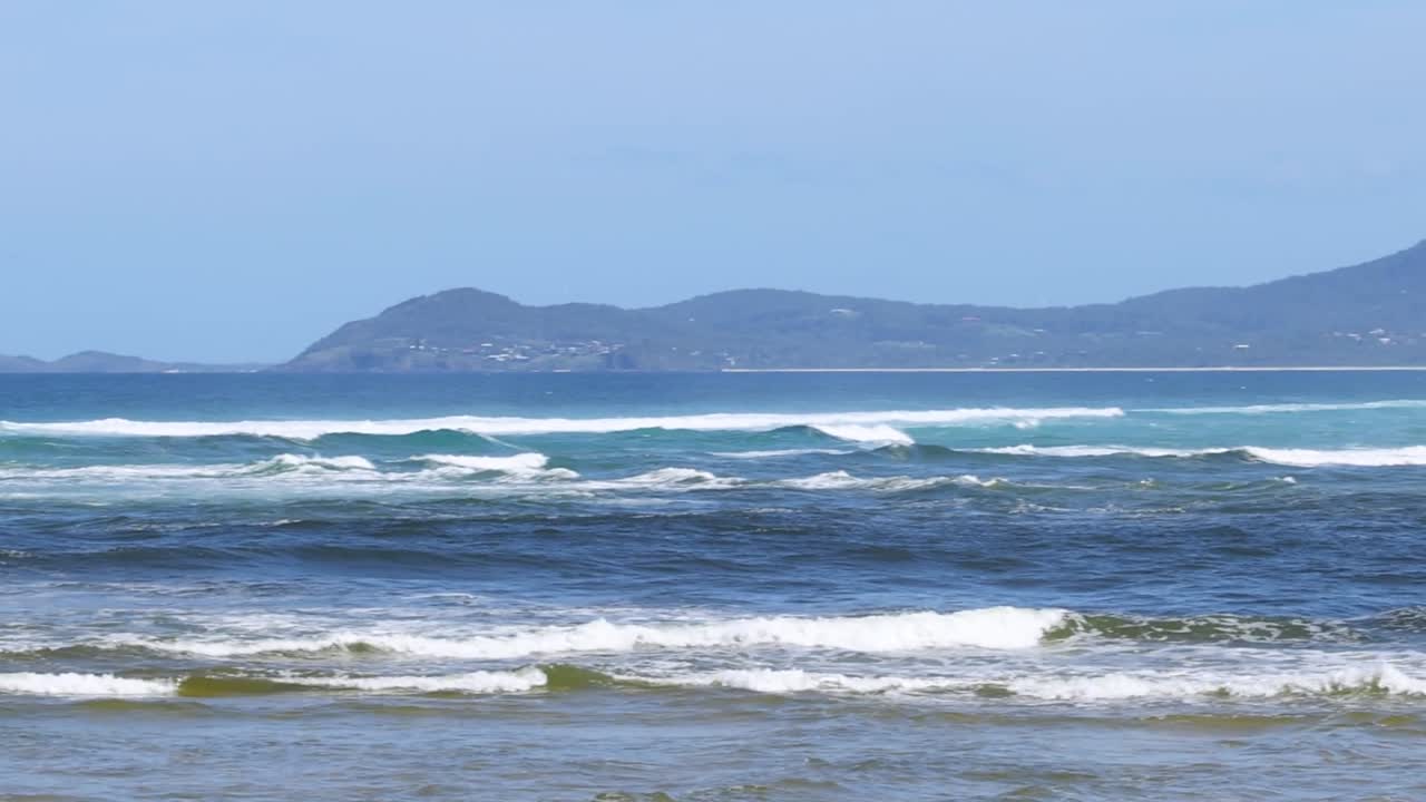 A serene view of ocean waves with distant hills and a clear blue sky.