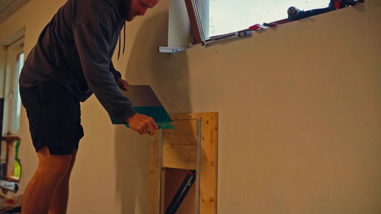 Un uomo sta lavorando all'interno, posizionando un pannello su una cornice di legno vicino al muro - Inquadratura fissa.